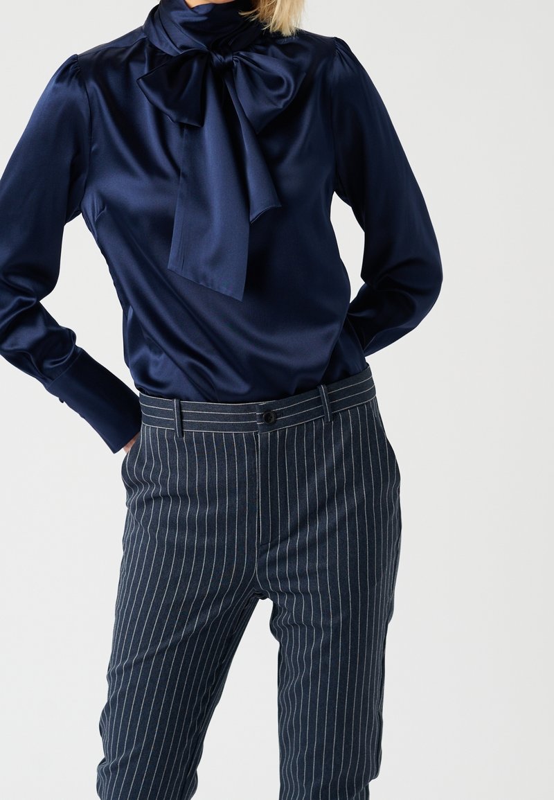 Blusa di raso blu navy con un grande colletto a fiocco, abbinata a pantaloni blu navy a righe con vita standard e chiusura a bottone.