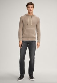 Hoodie beige a maglia con un motivo testurizzato, dotato di tasca frontale e cordini. Abbinato a jeans scuri e sneakers nere.