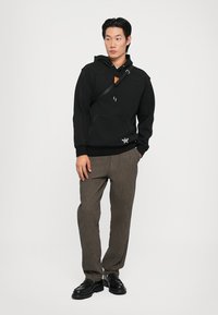 Black hooded sweatshirt with contrasting stitching, kangaroo pocket, and logo detail, paired with striped brown trousers and black shoes.