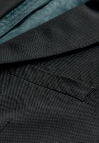 Close-up van de gestructureerde donkergrijze zak van een colbert en gedeeltelijk zichtbare blauwe, met patroon bedrukte binnenvoering.