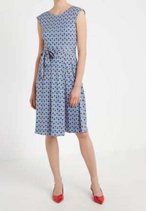 Femme portant une robe bleue sans manches longueur genou avec un motif géométrique et une ceinture nouée, associée à des talons rouges pointus.
