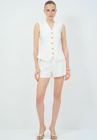 Chaleco sin mangas blanco con botones dorados, combinado con pantalones cortos blancos. Tejido suave, corte ajustado y diseño minimalista. Calzado con detalles dorados.
