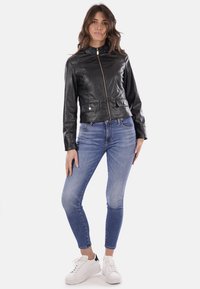 Giubbotto cropped in pelle nera con colletto alto, zipper frontale e due tasche laterali, abbinato a jeans skinny blu e sneakers bianche.