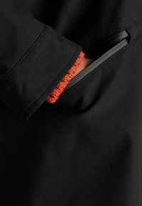 Strat exterior negru cu o textură netedă, dotat cu un manșon tricotat portocaliu și o deschidere pentru buzunar cu un design de fermoar contrastant.