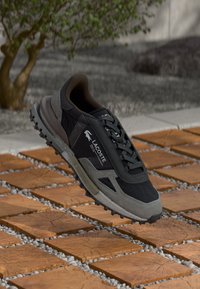 Lacoste sneaker in zwart en grijs, met een ontwerp van gemengde materialen, voorzien van een gestructureerde bovenkant, veters en een robuuste zool voor grip.