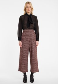 Blouse noire transparente avec un nœud papillon et des manches longues, associée à un pantalon bordeaux à jambes larges avec un motif géométrique, le tout surmonté de bottes noires à talons.