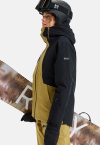 Black and gold outerwear, featuring a hood, zippered pockets, and contrast panels. Snowboard with a forest pattern in the background.