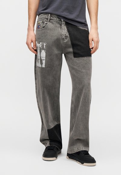 Φαρδιά γκρι τζιν με ανάμικτες υφές και μπαλώματα. Διαθέτουν πλαϊνή τσέπη με γραφικό σχέδιο. Συνδυάζονται με μαύρα αθλητικά παπούτσια.