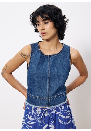 Top en denim court avec un col rond, sans manches, et des détails de couture apparente, porté par-dessus une jupe bleue à motifs floraux.