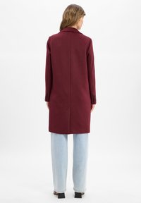 Cappotto in lana bordeaux con vestibilità sartoriale, maniche lunghe e collarino a dente, abbinato a jeans a gamba larga blu chiaro.