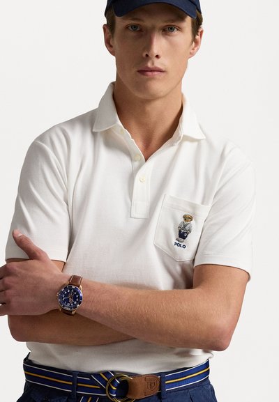 Hvid polo skjorte med en venstre brystlomme, der har et broderet logo. Båret med et navy bælte, navy bukser og et brunt ur med en blå urskive.