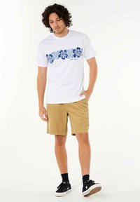 Hvid kortærmet t-shirt med blåt blomsterdesign og tekst; parret med beige fløjlshorts og sorte sneakers med hvide accenter.