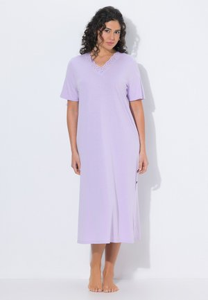 Frau mit lockigem Haar, die ein hellviolettes, kurzärmeliges, knöchellanges Kleid trägt und barfuß vor einem weißen Hintergrund steht.