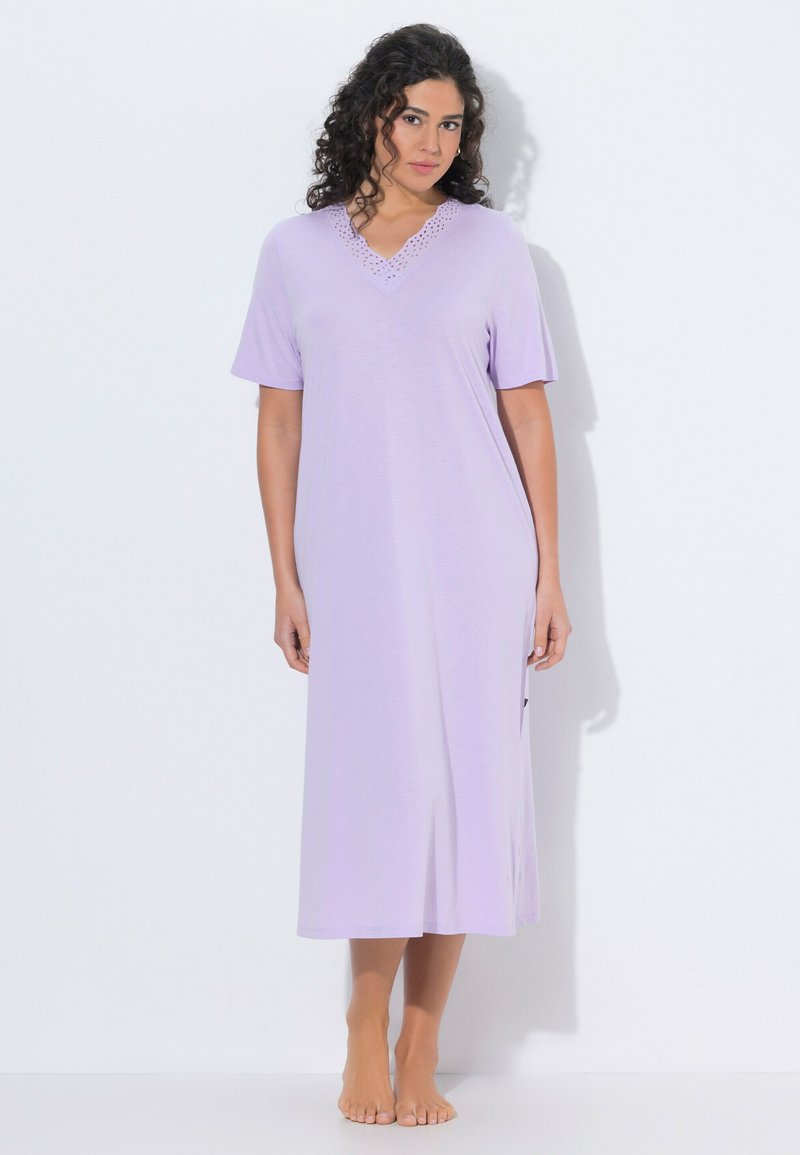 Mujer con cabello rizado, vestida con un vestido de manga corta, color púrpura claro, hasta el tobillo, de pie descalza sobre un fondo blanco.