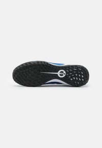 Zapatilla deportiva azul con una suela exterior de goma negra texturizada, que presenta un acento blanco en la entresuela y un diseño de logo en el talón.