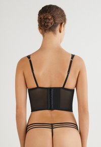 Svart underklädesset med en genomskinlig bodice i tyg, justerbara axelband och hak-och-öga-stängning, tillsammans med en matchande stringtrosa med remdetaljer.