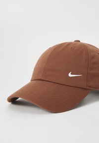 Καφέ καπέλο μπέιζμπολ από υφασμάτινη ύφανση με καμπύλο γείσο και λευκό λογότυπο Nike στο μπροστινό μέρος. Διαθέτει οπή εξαερισμού στην κορυφή.