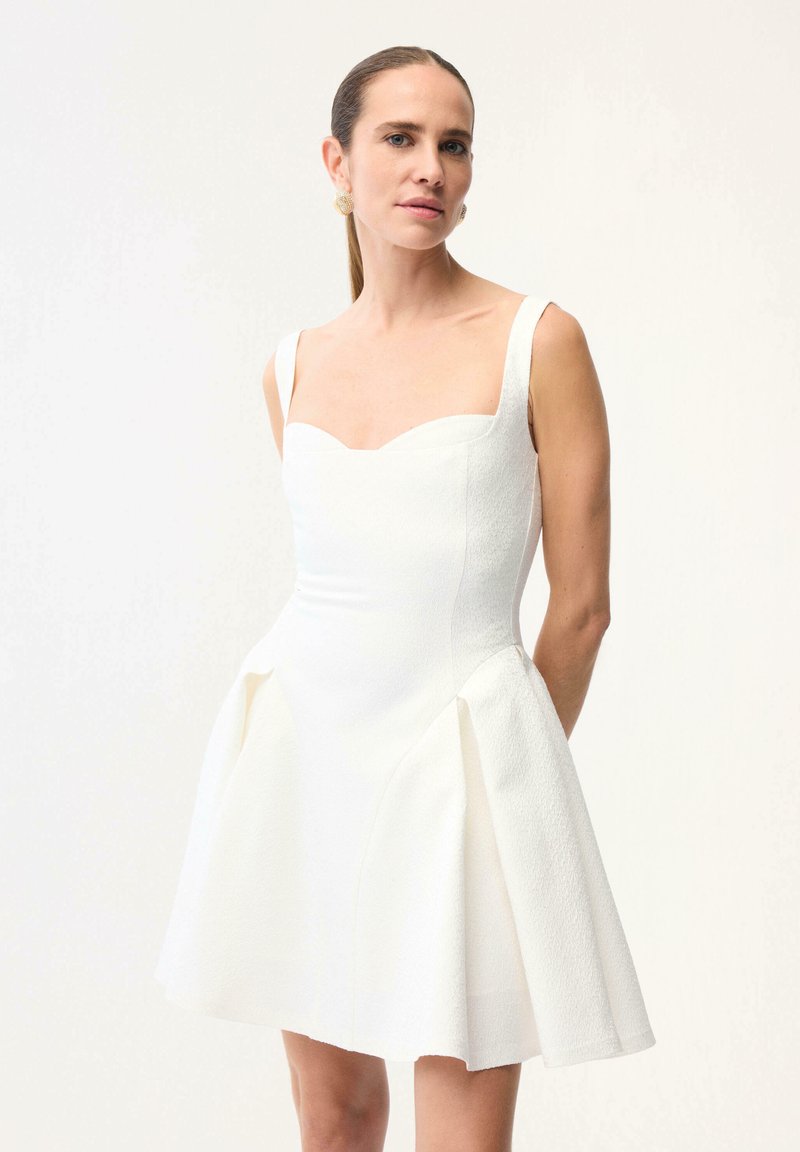 Femme aux cheveux raides tirés en arrière, portant une mini-robe blanche texturée sans manches avec un corsage ajusté et une jupe évasée, sur un fond uni.