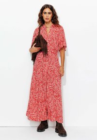 Robe maxi à motif paisley rouge avec des manches courtes et un devant à boutons. Associée à un sac bandoulière en cuir marron à franges et des bottes foncées.