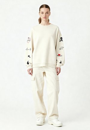 Felpa crema con stampe grafiche sulle maniche, abbinata a pantaloni cargo crema coordinati. Calzature sono sneakers bianche. Sfondo neutro.