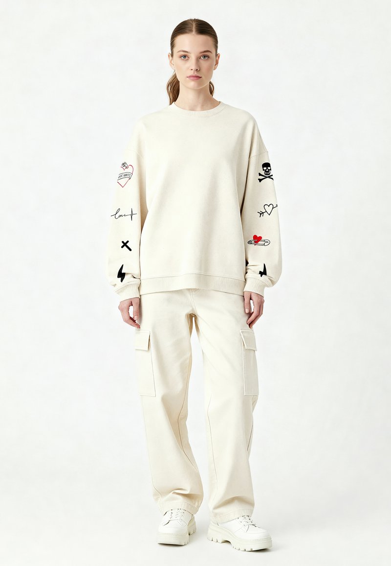 Felpa crema con stampe grafiche sulle maniche, abbinata a pantaloni cargo crema coordinati. Calzature sono sneakers bianche. Sfondo neutro.