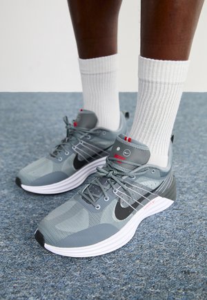 Person trägt graue Nike Laufschuhe und weiße, gerippte Crew-Socken und steht auf blauem Teppichboden.