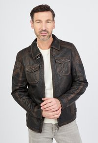 Braune Lederjacke mit strukturierten Einsätzen, doppelten Brusttaschen und kontrastierenden Nähten; über einem hellen Hemd getragen. Modell posiert mit verschränkten Händen.