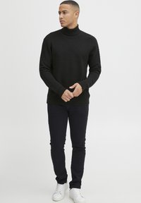 Maglione a collo alto nero con texture, con polsini e orlo a costine, abbinato a jeans scuri e sneakers bianche, su uno sfondo neutro.