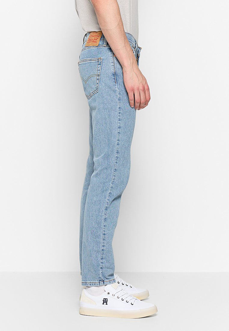 Jeans de mezclilla azul claro con un corte recto, que presentan bolsillos traseros y un parche de cuero marrón claro en la cintura. Llevados con zapatillas blancas.