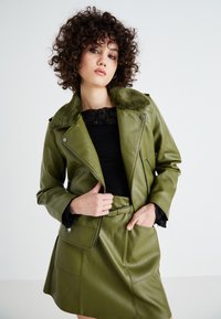 Green faux leather biker jacket with a soft fur collar, paired with a black lace top and green skirt featuring pockets and belt detail.