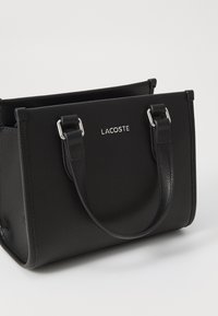 Sac fourre-tout en cuir noir avec surface texturée, doubles poignées supérieures et logo Lacoste métallique à l'avant. Forme rectangulaire, design structuré.