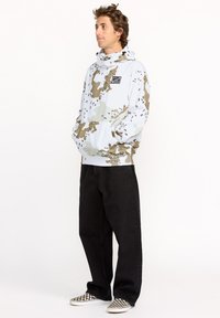 Sudadera con capucha en blanco con un patrón de camuflaje en tonos marrones y verdes; cuenta con un bolsillo delantero y puños acanalados. Combinada con pantalones negros.