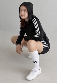 Schwarzer Kapuzenpullover mit weißen Streifen, kombiniert mit schwarzen Shorts und weißen knielangen Socken. Weiße Sportschuhe runden das Outfit ab.