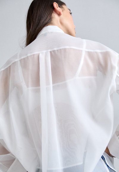 Transparent hvid bluse med krave, der har en løs pasform, lange ærmer og fine sydetaljer langs ryggen og manchetterne.