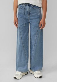 Weite Jeans in hellblauem Denim, mit hoher Taille, sichtbaren Nähten und einem schmalen Schnitt am Saum. Kombiniert mit weißen Turnschuhen.