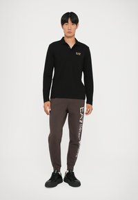 Juodas ilgarankovis polo marškinėliai su EA7 logotipu, derinami su rudais sportiniais šortais su balta užrašu. Modelis vilki juodus sportbačius, stovi prieš baltą foną.