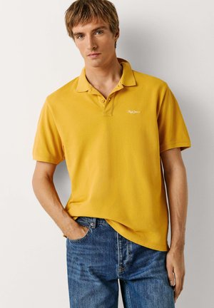 Noor mees, kes kannab sinepikollast lühikeste varrukatega polo särk ja siniseid teksaseid, seisab ühe käega püksitaskus.