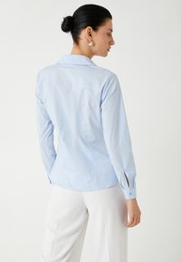 Camisa de botones de manga larga en azul claro con cuello estructurado. Presenta un dobladillo recto y puños abotonados con aberturas. Textura de tela suave.