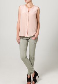Blusa sin mangas de color rosa claro con un botón en la parte delantera, combinada con pantalones ajustados verdes y sandalias de tacón negro. Detalle de collar dorado visible.