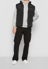 Gilet imbottito nero con cappuccio, sopra un felpa grigia, abbinato a pantaloni cargo neri e sneakers bianche. Texture liscia e lucida, con zip frontale.