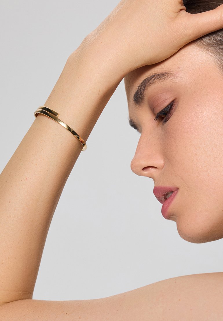 Złoty bransoletka na nadgarstek z eleganckim, wypolerowanym wzornictwem. Posiada gładką powierzchnię i cienki, zakrzywiony kształt, noszona na nadgarstku.
