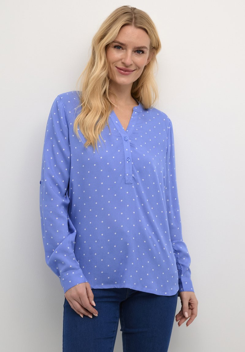 Blouse à manches longues bleu clair avec un décolleté en V, ornée de pois blancs et d'une patte de boutonnage. Tissu doux avec une coupe décontractée.