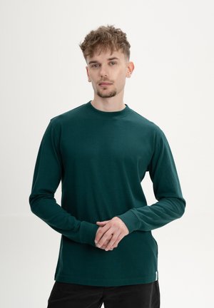 Jonge man met kort krullend haar, gekleed in een donkergroen shirt met lange mouwen, staand met gevouwen handen tegen een effen witte achtergrond.