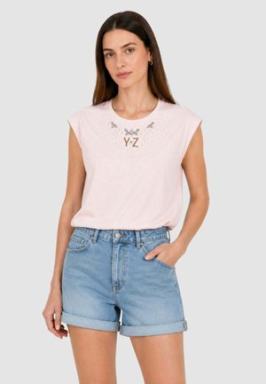 Donna con lunghi capelli scuri che indossa una canotta rosa con la scritta "Y·Z" e pantaloncini di denim azzurri chiari risvoltati, in piedi con una mano in tasca.