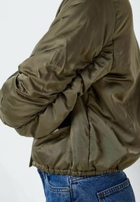 Olivgrön puffjacka med glansig yta, ribbad nederkant och kraftiga ärmar. Har sidfickor och en avslappnad passform. Kombineras med denimjeans.