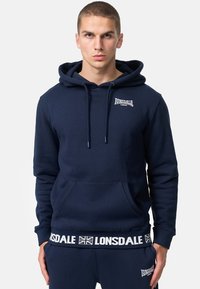 Tengerészkék kapucnis pulóver, húzózsinórral, elülső zsebbel, és Lonsdale logóval a mellkasán és az derékrészén. Puha anyagból készült, sima tapintással.