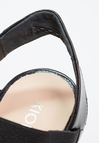 Sandale noire présentant un matériau texturé, un design à dos ouvert, et un talon brillant accentué avec une semelle intérieure beige. Logo "Kio" visible.