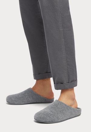 Graue Hausschuhe mit offenem Rücken, weichem Material und abgerundetem Design, kombiniert mit gerollten grauen Hosen, die einen lässigen Look zeigen.