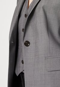 Blazer gris sur mesure avec un tissu texturé, une fermeture à un bouton, des revers crantés et une poche poitrine.