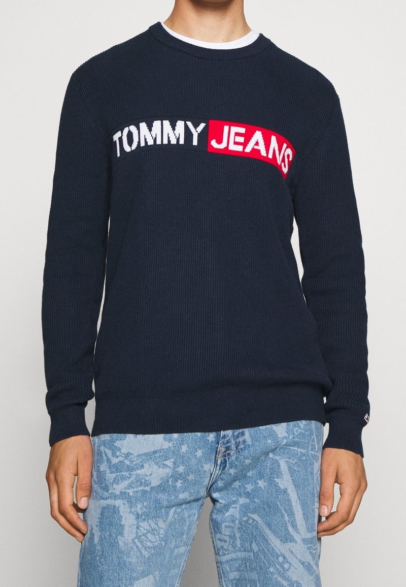 Granatowy sweter dziany o strukturalnym splocie, z białym i czerwonym logo „TOMMY JEANS” na piersi, z okrągłym dekoltem i kontrastowymi mankietami.